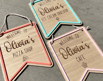 May include: Wooden pennant-shaped signs with the text "Welcome to Olivia's" and the business type: Pizza Shop, Ice Cream Parlour, and Cafe. Each sign has a colored border and a rope for hanging. The signs are made of light-colored wood.