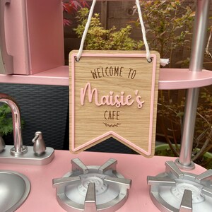 Puede incluir: Una cocina de juguete de madera rosa con un letrero que dice "Bienvenido al Caf&eacute; de Maisie". El letrero est&aacute; hecho de madera y tiene un borde rosa. La cocina tiene un grifo plateado y dos quemadores de estufa plateados.