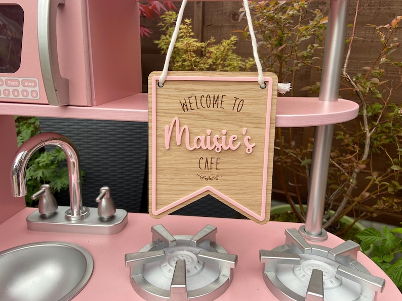 Puede incluir: Una cocina de juguete de madera rosa con un letrero que dice "Bienvenido al Caf&eacute; de Maisie". El letrero est&aacute; hecho de madera y tiene un borde rosa. La cocina tiene un grifo plateado y dos quemadores de estufa plateados.