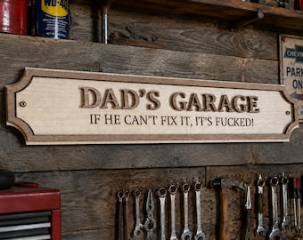 May include: A rectangular wooden sign with decorative edges featuring the text DAD'S GARAGE and the phrase IF HE CAN'T FIX IT, IT'S FUCKED! in dark brown raised lettering against a light wood background, secured with two visible metal screws.