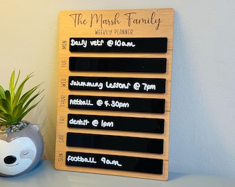 May include: Wooden weekly planner with the text "The Marsh Family" at the top. It has black rectangular sections for each day of the week, with handwritten appointments like "Daily vets @ 10am" and "Swimming lessons @ 7pm".