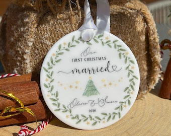 May include: A round white ceramic ornament with a white ribbon hanger. The design features a green leaf wreath, gold stars, and a green Christmas tree. Text reads: Our FIRST CHRISTMAS married, Oliver & Simone, 2026. The ornament has a smooth finish.