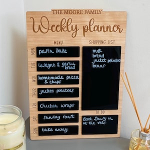 May include: A wooden weekly planner with a chalkboard surface. The planner has sections for a menu, shopping list, and to-do list. The menu section lists meals for each day of the week, including pasta bake, lasagna, homemade pizza, jacket potatoes, chicken wraps, Sunday roast, and take away. The shopping list includes milk, bread, jacket potatoes, and beans. The to-do list includes "Book Daisy in at the vets".