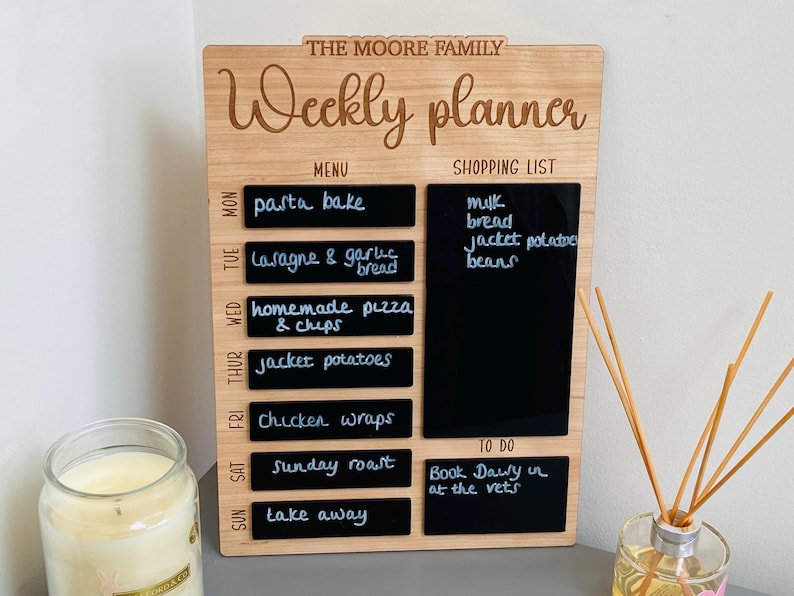 May include: A wooden weekly planner with a chalkboard surface. The planner has sections for a menu, shopping list, and to-do list. The menu section lists meals for each day of the week, including pasta bake, lasagna, homemade pizza, jacket potatoes, chicken wraps, Sunday roast, and take away. The shopping list includes milk, bread, jacket potatoes, and beans. The to-do list includes "Book Daisy in at the vets".