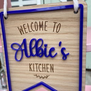 Puede incluir: Un letrero de madera con un borde azul y el texto "Bienvenido a la cocina de Albie" en una fuente cursiva.