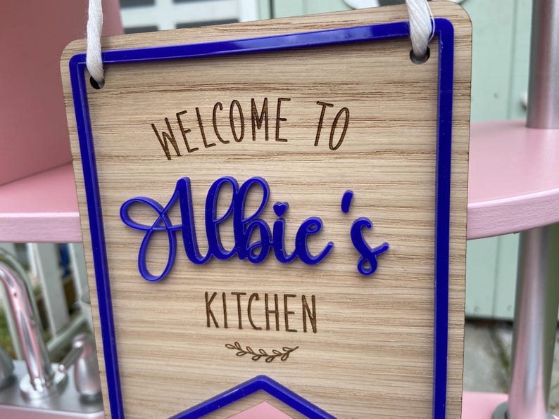 Puede incluir: Un letrero de madera con un borde azul y el texto "Bienvenido a la cocina de Albie" en una fuente cursiva.