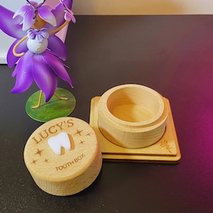 May include: A wooden tooth box with the name "LUCY'S" and a tooth graphic engraved on the lid. The box is open and the lid is sitting next to it. The box is on a dark surface.