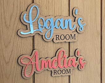 May include: Two wooden room signs with names in cursive script. The top sign reads "Logan's ROOM" in blue and brown. The bottom sign reads "Amelia's ROOM" in pink and brown. The signs are mounted on a wooden door.