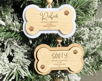 May include: Two wooden dog bone ornaments with the names "Rufus" and "Sooty" engraved on them. The ornaments are decorated with paw prints and the text "My first Christmas in my forever home 2024".