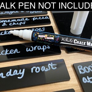 May include: A black chalkboard with a white chalk marker. The chalkboard is divided into seven sections, each labeled with a day of the week. The sections are filled with handwritten text, such as "lasagne", "homemade pizza", "chicken wraps", and "Sunday roast". The text "CHALK PEN NOT INCLUDED!" is printed at the top of the chalkboard.