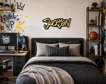 May include: A custom wall sign featuring the name Jackson in a bold, yellow and black graffiti-style font. The sign is mounted on a plain wall above a bed with dark gray bedding, pillows, and a textured throw blanket in a bedroom setting.