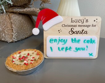 May include: A wooden sign with a Santa hat on top. The sign says "Lucy's Christmas message for Santa" and has a white dry-erase board attached to it. The board says "enjoy the cake I left you!" There is a small pie with red and white sprinkles on top in the foreground.