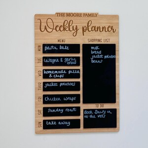 May include: A wooden weekly planner with a chalkboard surface. The planner has sections for a menu, shopping list, and to-do list. The menu section lists meals for each day of the week, such as pasta bake, lasagna, and chicken wraps. The shopping list includes items like milk, bread, and beans. The to-do list has one item: "Book Dawy in at the vets."