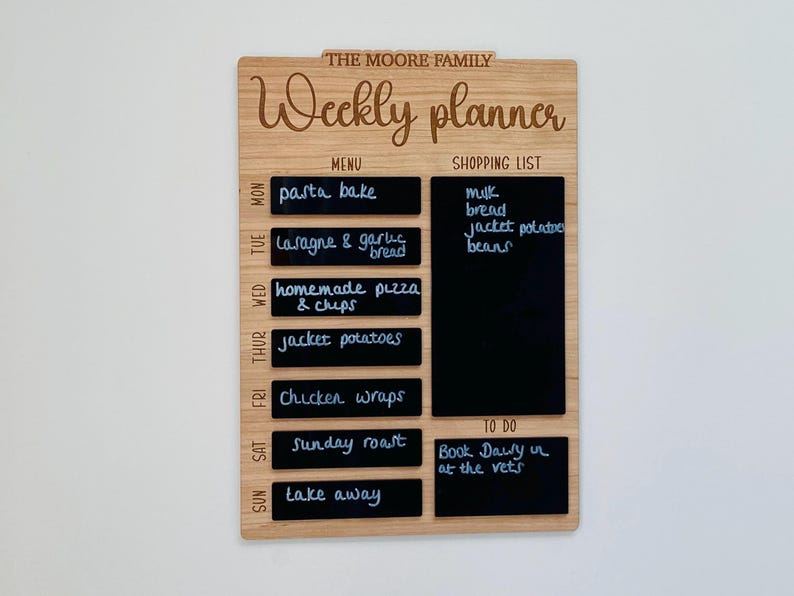 May include: A wooden weekly planner with a chalkboard surface. The planner has sections for a menu, shopping list, and to-do list. The menu section lists meals for each day of the week, such as pasta bake, lasagna, and chicken wraps. The shopping list includes items like milk, bread, and beans. The to-do list has one item: "Book Dawy in at the vets."