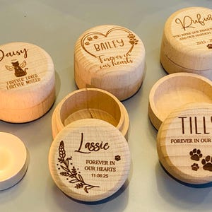 May include: A collection of wooden memorial boxes for pets, each engraved with a name and a message such as 'Forever in our hearts'. The boxes are round with lids and feature paw prints and floral designs. A lit candle is visible.