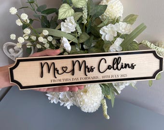 May include: A wooden sign with the words "Mr & Mrs Collins" in black script, a heart symbol, and the date "10th July 2025". The sign has a black border and is set against a floral backdrop.