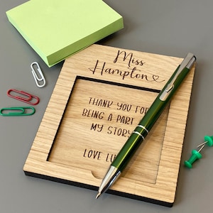 May include: A wooden picture frame with the text "Miss Hampton" and "Thank you for being a part of my story". A green pen, green sticky notes, and paper clips are also present.