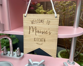 May include: A wooden sign with the text "Welcome to Maisie's Kitchen" in a cursive font. The sign is hanging from a white string and is attached to a pink shelf.