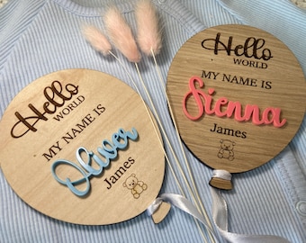 May include: Two wooden balloon ornaments with the text "Hello World My Name Is" and the names "Oliver" and "Sienna" in blue and pink lettering.  Each balloon has a small teddy bear graphic and a white ribbon.