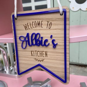 Puede incluir: Un letrero de madera con un borde azul y el texto "Bienvenido a la cocina de Albie" en una fuente decorativa.