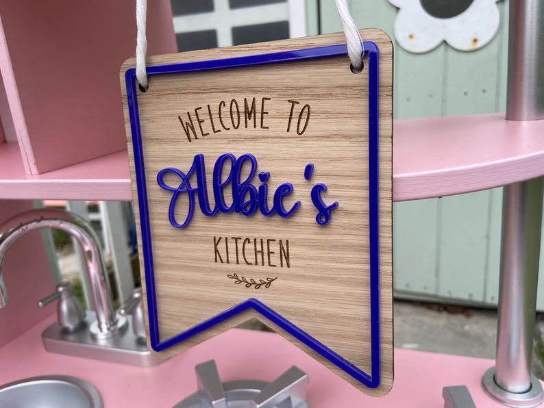 Puede incluir: Un letrero de madera con un borde azul y el texto "Bienvenido a la cocina de Albie" en una fuente decorativa.