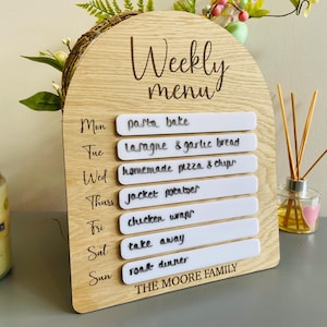 May include: A wooden weekly menu board with white removable labels. The board is decorated with a wood grain pattern and the text "Weekly menu" is engraved on the top. The labels list the days of the week and the meals planned for each day. The text on the labels includes "pasta bake", "lasagna & garlic bread", "homemade pizza & chips", "jacket potatoes", "chicken wraps", "take away", and "roast dinner". The bottom of the board has the text "THE MOORE FAMILY".