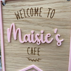 Puede incluir: Un letrero de madera con un borde rosa y el texto "Bienvenido a Maisie's Cafe" en letras negras. El nombre "Maisie's" est&aacute; en letras rosas en relieve con dos peque&ntilde;os corazones encima.