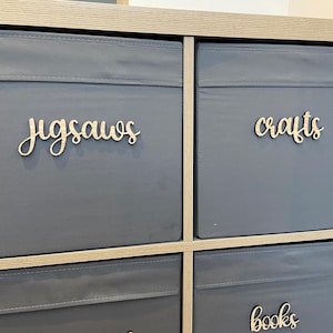 May include: A set of three gray fabric storage bins with wooden labels. The labels read "jigsaws", "crafts", and "books".