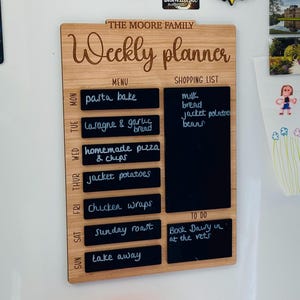 May include: A wooden weekly planner with a chalkboard surface. The planner has a menu section with a list of meals for each day of the week, a shopping list section, and a to-do list section. The planner is labeled "The Moore Family Weekly Planner".