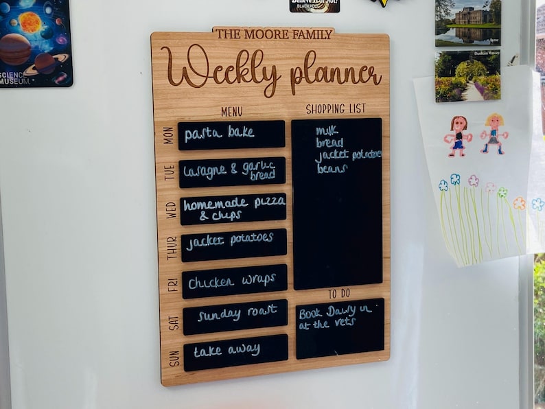 May include: A wooden weekly planner with a chalkboard surface. The planner has a menu section with a list of meals for each day of the week, a shopping list section, and a to-do list section. The planner is labeled "The Moore Family Weekly Planner".