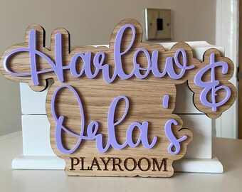 May include: Wooden sign with the names "Harlow & Orla's" in lavender script, above the word "PLAYROOM" in a block font. The sign has a light wood grain background and is mounted on a white surface.