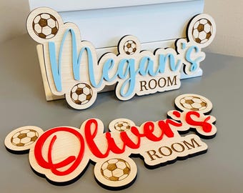 May include: Wooden room signs with soccer ball accents. One sign reads "Megan's ROOM" in blue and the other reads "Oliver's ROOM" in red. The signs are made of light-colored wood with soccer ball designs.