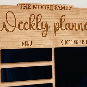 May include: A wooden weekly planner with the text "The Moore Family Weekly Planner" at the top. The planner has sections for "Menu" and "Shopping List" with blank spaces for writing.