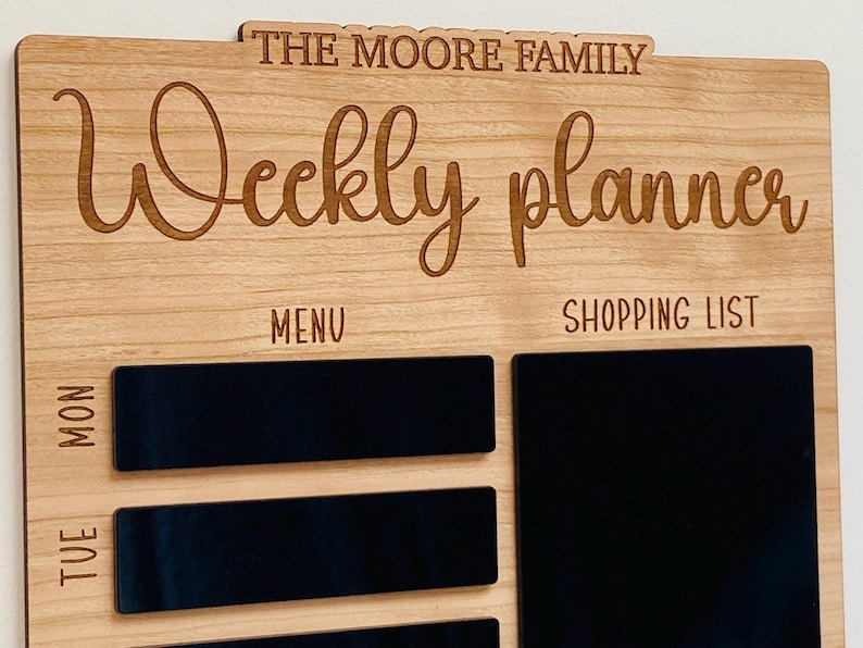 May include: A wooden weekly planner with the text "The Moore Family Weekly Planner" at the top. The planner has sections for "Menu" and "Shopping List" with blank spaces for writing.