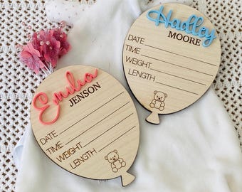 May include: Two wooden balloon-shaped baby announcement signs. One is pink with the name "Emilia Jenson" and the other is blue with the name "Hadley Moore". Both signs have spaces to record the baby's date, time, weight, and length.