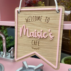Puede incluir: Un letrero de madera rosa con un borde rosa y el texto "Bienvenido al Caf&eacute; de Maisie" en letras marrones. El letrero est&aacute; colgado de una cuerda blanca.