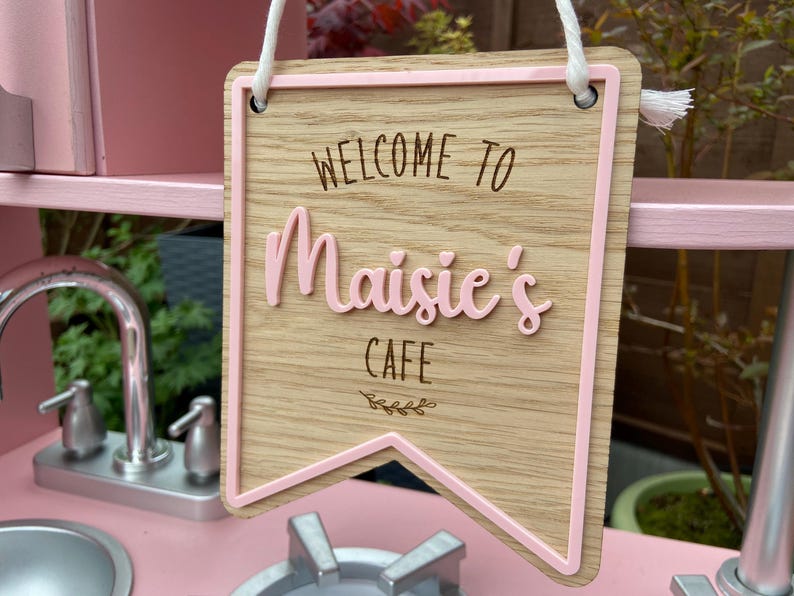 Puede incluir: Un letrero de madera rosa con un borde rosa y el texto "Bienvenido al Caf&eacute; de Maisie" en letras marrones. El letrero est&aacute; colgado de una cuerda blanca.