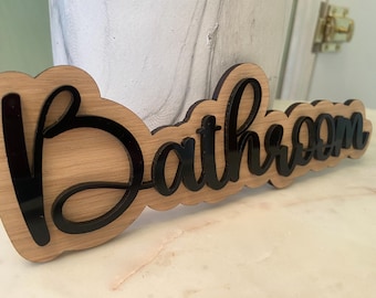 May include: Wooden "Bathroom" sign with black cursive lettering. The sign is layered, with a light brown wood base and black acrylic letters. The sign is designed to be mounted on a wall or door.