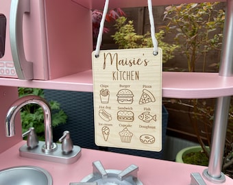 May include: A wooden sign with the text "Maisie's Kitchen" and a list of food items, including chips, burger, pizza, hot dog, sandwich, fish, ice cream, cupcake, and doughnut. The sign is hanging on a pink play kitchen.