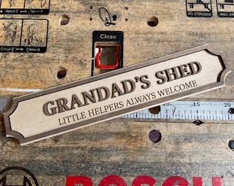 May include: A rectangular wooden sign with decorative notched corners, featuring the raised dark brown text "GRANDAD'S SHED" and the engraved text "LITTLE HELPERS ALWAYS WELCOME" in a lighter wood finish, positioned above a metal ruler.