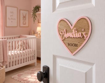 May include: A heart-shaped wooden door sign with a light pink border. The sign features the text Amelia's in raised pink script lettering and the word ROOM in dark brown block letters centered on a natural wood grain background.