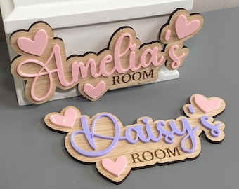 May include: Two wooden wall signs with light wood grain finish. One sign features the pink acrylic text Amelia's ROOM with pink hearts. The other sign features the purple acrylic text Daisy's ROOM with pink hearts. Both have scalloped edges.