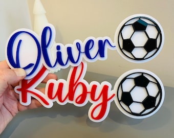 May include: A set of personalized soccer-themed decorations. The design includes the names "Oliver" in blue and "Ruby" in red, along with two soccer ball cutouts. The names are in a cursive font, and the soccer balls are black and white.