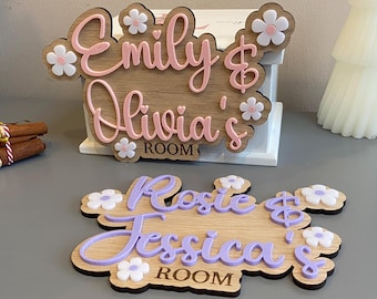 May include: Wooden room signs with names in cursive script. One sign reads "Emily & Olivia's ROOM" in pink, and the other reads "Rosie & Jessica's ROOM" in purple. Both signs have flower accents.