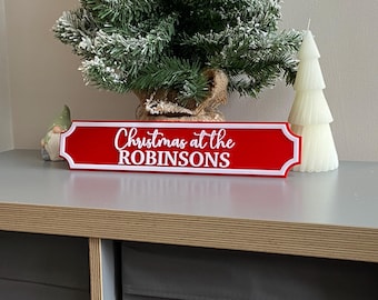 May include: A red sign with white trim and the text "Christmas at the ROBINSONS" in white script. A small Christmas tree and a white tree-shaped candle are in the background. A small gnome figurine is on the left.