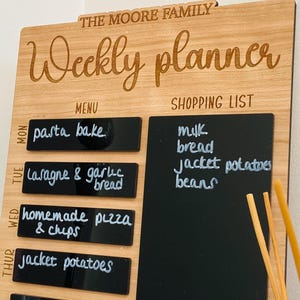 May include: A wooden weekly planner with a chalkboard shopping list and a menu section. The menu section has a list of meals for each day of the week, including pasta bake, lasagna, garlic bread, homemade pizza, and chips. The shopping list includes milk, bread, jacket potatoes, and beans.