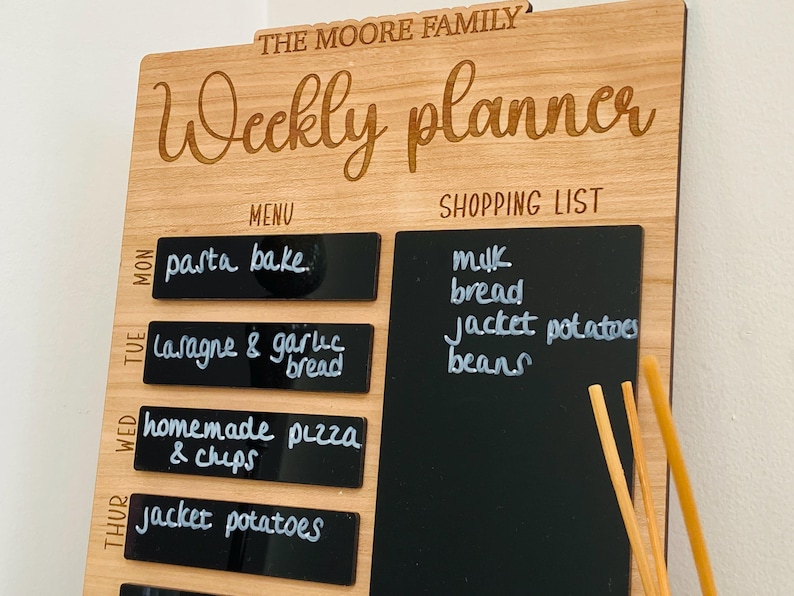 May include: A wooden weekly planner with a chalkboard shopping list and a menu section. The menu section has a list of meals for each day of the week, including pasta bake, lasagna, garlic bread, homemade pizza, and chips. The shopping list includes milk, bread, jacket potatoes, and beans.