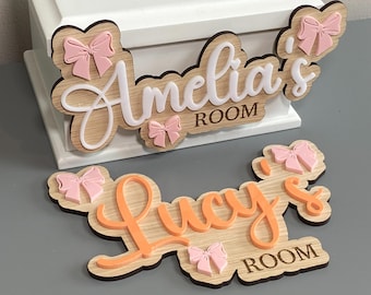 May include: Two custom wooden signs with light wood grain finish, featuring raised script lettering and small pink bows. One sign displays the text Amelia's ROOM in white, and the other displays the text Lucy's ROOM in orange, both with scalloped edges.