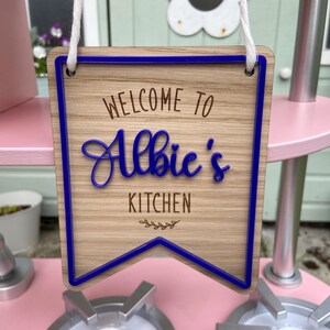 Puede incluir: Un letrero de madera con ribete azul que dice "Bienvenido a la cocina de Albie". El letrero est&aacute; colgado de una cuerda blanca.
