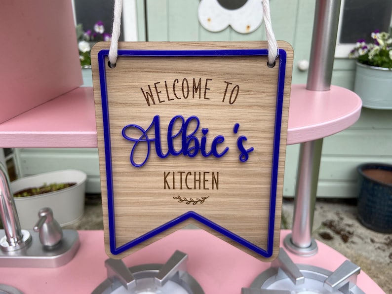 Puede incluir: Un letrero de madera con ribete azul que dice "Bienvenido a la cocina de Albie". El letrero est&aacute; colgado de una cuerda blanca.